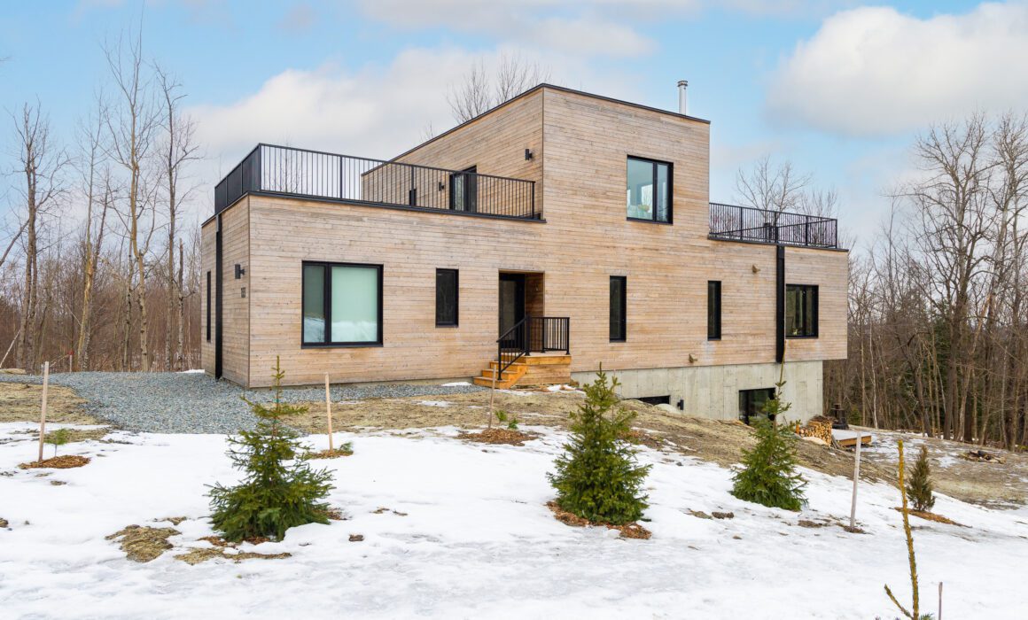 Façade d’une maison contemporaine en bois et béton, implantée en pleine nature, avec escaliers d’entrée, terrasses et jeunes conifères en premier plan.