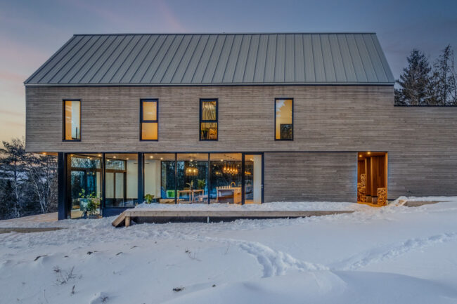 Façade arrière épurée où l’architecture contemporaine embrasse la nature : lignes sobres, matériaux nobles et vastes ouvertures sur l’hiver québécois.