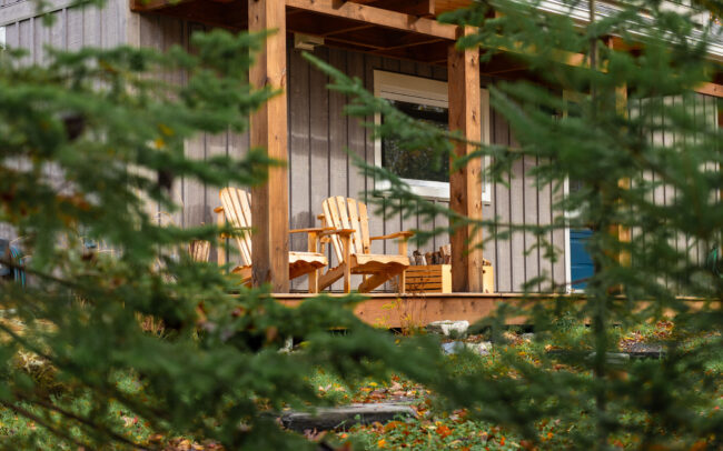 Vue intimiste à travers les conifères sur la terrasse en bois, où deux fauteuils Adirondack invitent à la détente en pleine nature.