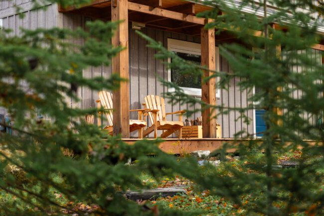 Vue intimiste à travers les conifères sur la terrasse en bois, où deux fauteuils Adirondack invitent à la détente en pleine nature.