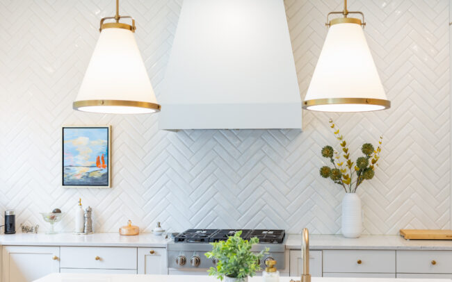 Vue rapprochée d’un mur en céramique à chevrons blanc cassé avec hotte centrale, luminaires suspendus dorés, et comptoir de cuisine décoré de vases et accessoires
