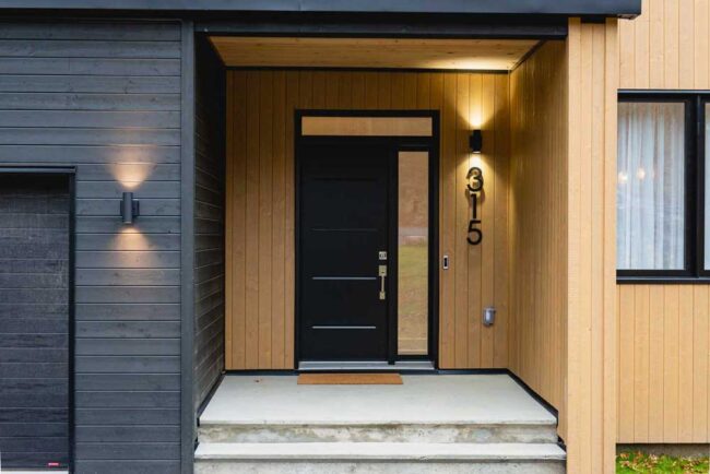 Entrée chaleureuse et contrastée avec revêtement en bois naturel, porte noire moderne et éclairage architectural mettant en valeur l'adresse.